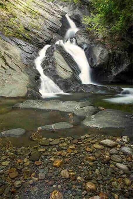 Serene waterfall cascading gracefully over smooth, grey rock formations into a clear, shallow pool filled with pebbles, surrounded by lush green foliage.