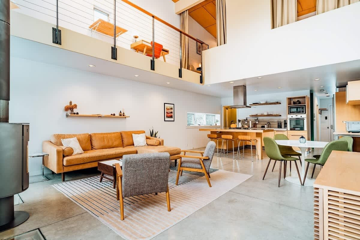 Open-concept living space with high ceilings, concrete floors, and a mezzanine level, featuring a mustard yellow sofa, a modern wood-burning stove, and an adjacent kitchen with a large island.