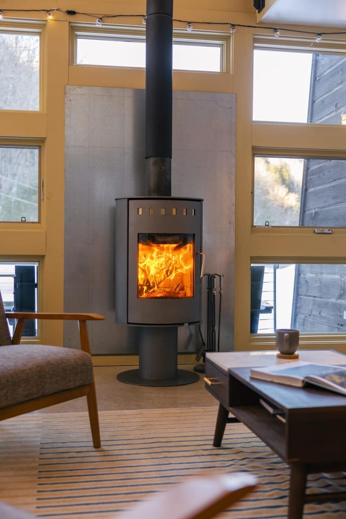 A cozy modern living room features a dark gray wood-burning stove with a visible fire, set against a light gray panel and large windows. A striped rug, a mid-century modern armchair, and a coffee table with a mug and book complete the inviting space.