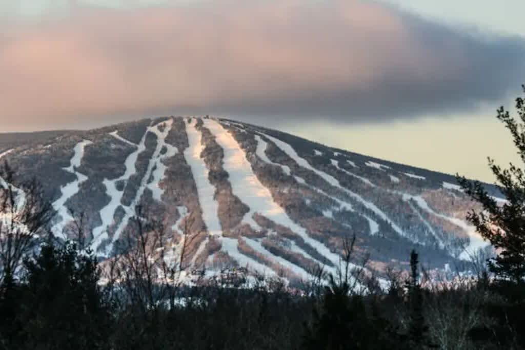 A vast snow-covered mountain with numerous ski trails visible, illuminated by the soft light of a cloudy sunset sky. Dark evergreen trees line the base of the mountain.