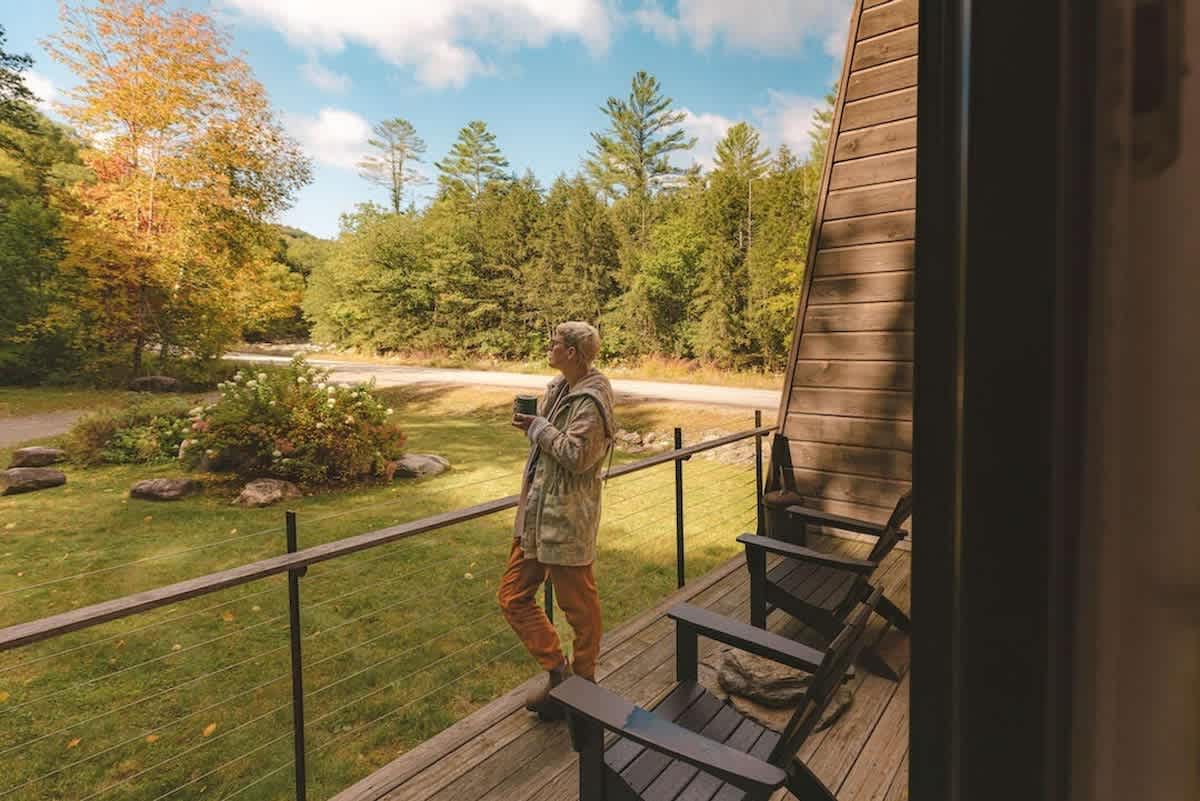 A person stands on a wooden balcony overlooking a lush green yard and a road, surrounded by trees with autumn foliage. Black Adirondack chairs are visible on the deck, suggesting a relaxing outdoor space.