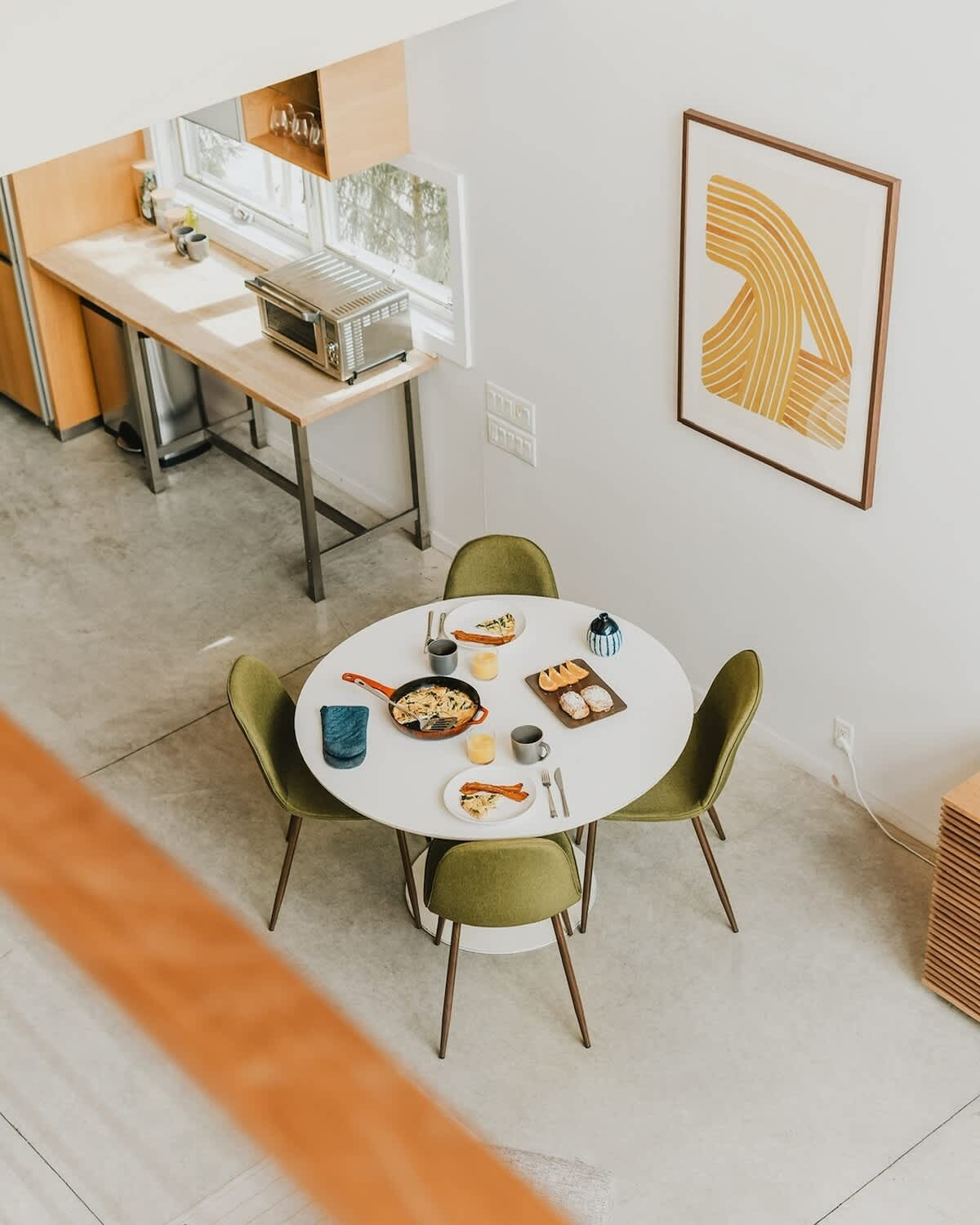 Overhead view of a modern dining area featuring a white oval table set with breakfast, surrounded by four green upholstered chairs. The space opens to a kitchen area with light wood countertops and a toaster oven.