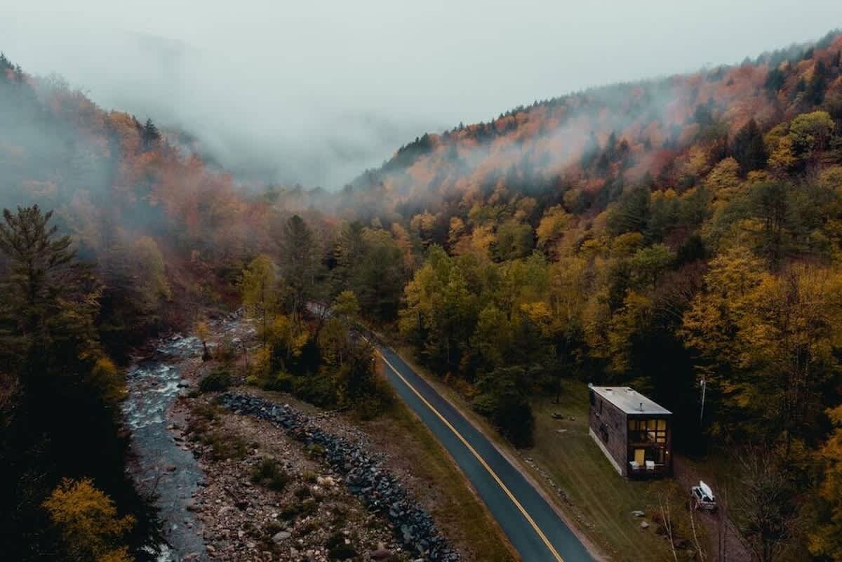 An aerial view captures a modern cabin with dark wood siding and large windows nestled beside a winding road and a river, surrounded by a vibrant autumn forest. The misty mountains in the background complete the picturesque, secluded setting.