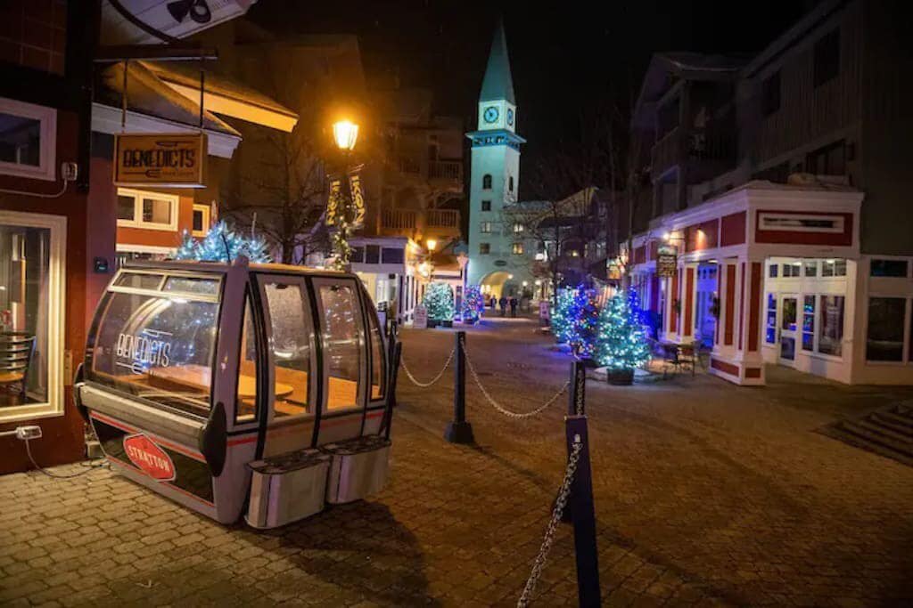 A festive village street at night, adorned with blue and white Christmas lights on trees and buildings, featuring a gondola car in the foreground and a clock tower in the background.