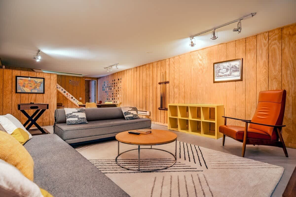 Another view of the wood-paneled game room, featuring a long gray sofa with throw pillows, a round wooden coffee table, and a geometric rug. An orange leather armchair, a yellow cubby shelf, and a foosball table are also visible, creating a cozy entertainment space.