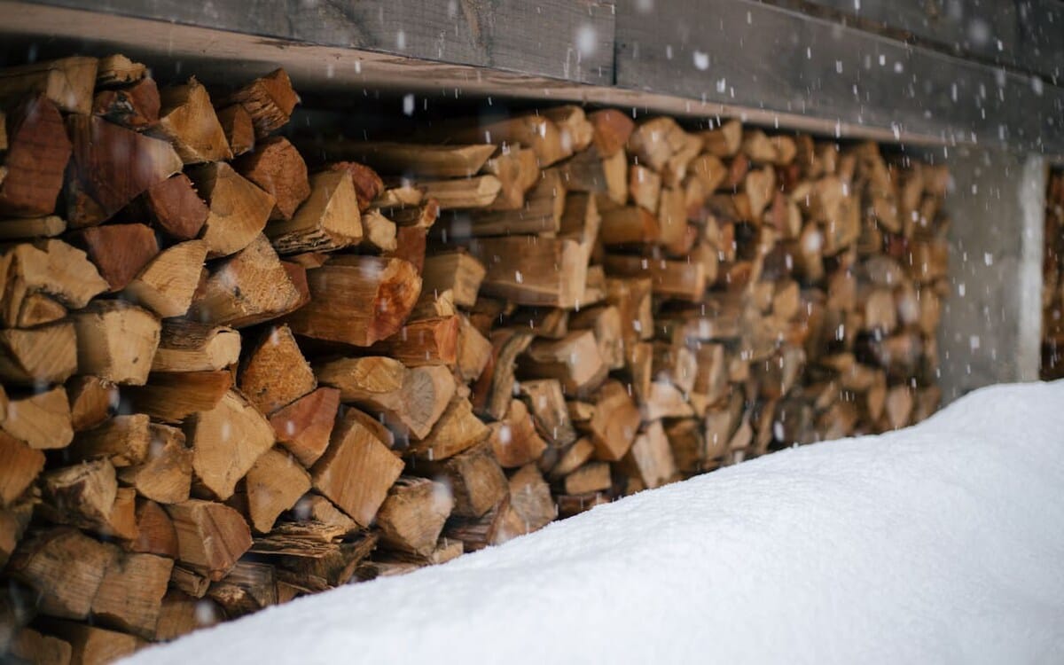 A neatly stacked pile of firewood is visible under a shelter, partially covered by fresh snow falling gently. This image highlights the practical amenities for a cozy stay in a winter environment.