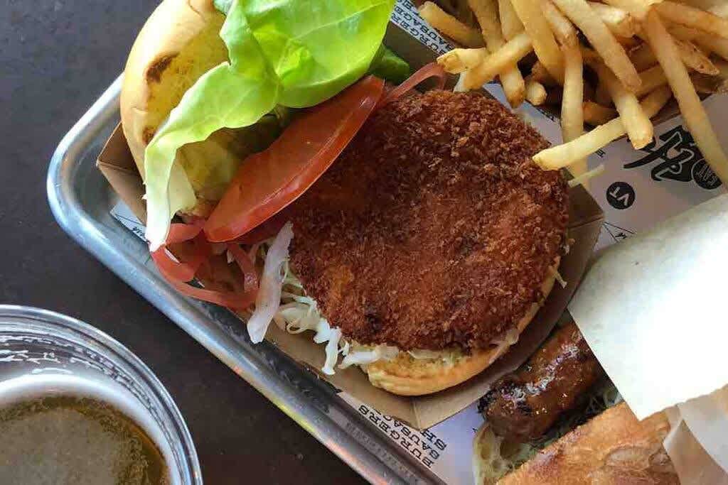 Close-up of a crispy fried burger with lettuce, tomato, and shredded cabbage on a bun, served with a side of golden french fries and a glass of beer on a metal tray.