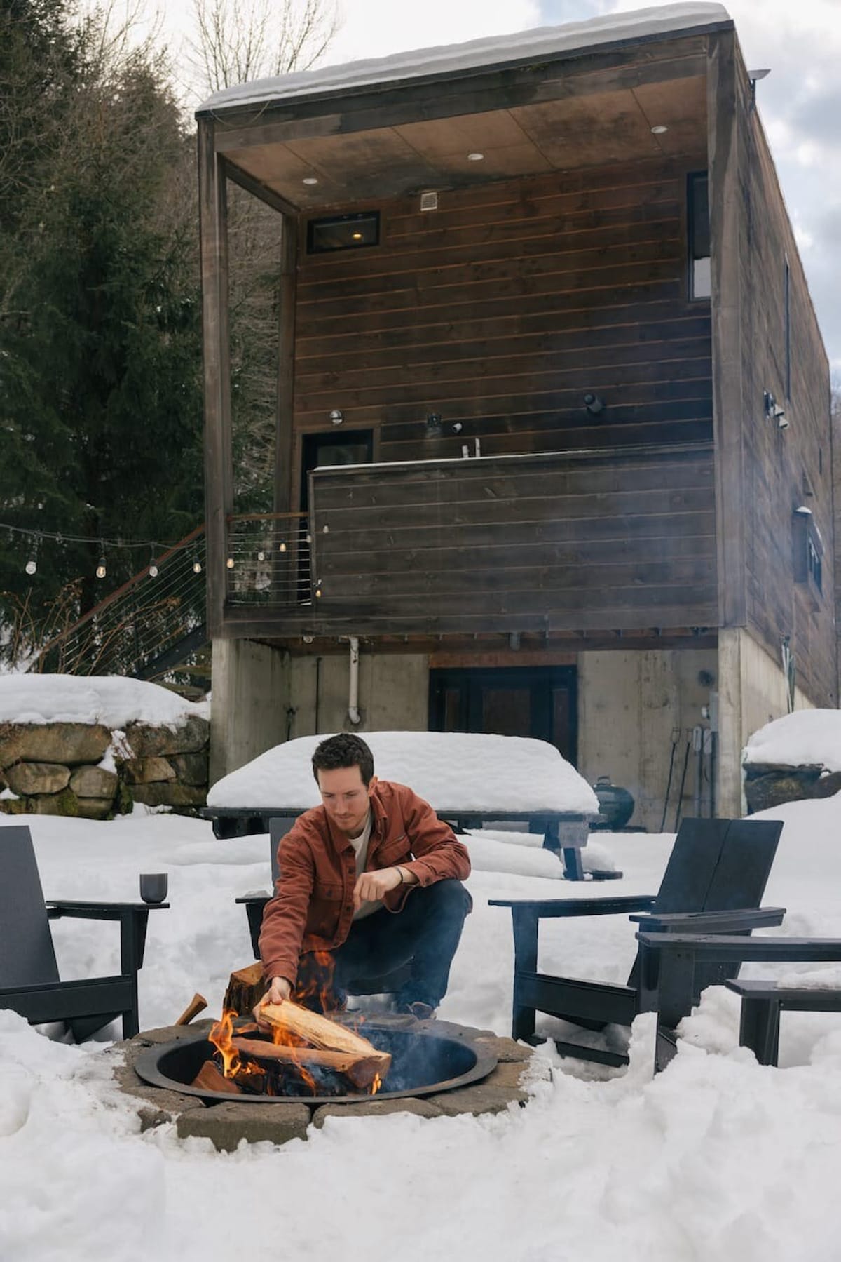 A man tends to a crackling fire in a stone fire pit, surrounded by snow-covered ground and black Adirondack chairs, with a modern cabin visible in the background. The scene evokes a cozy winter outdoor experience.