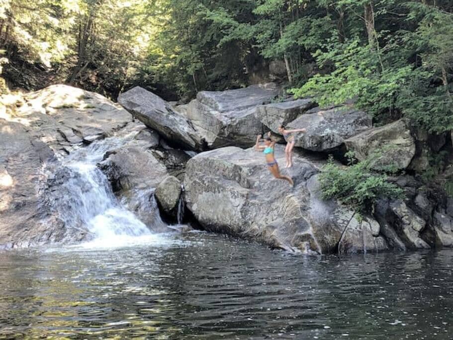 Two people jumping from large rocks into a natural swimming hole with dark water, next to a small waterfall, surrounded by dense green trees and large boulders.