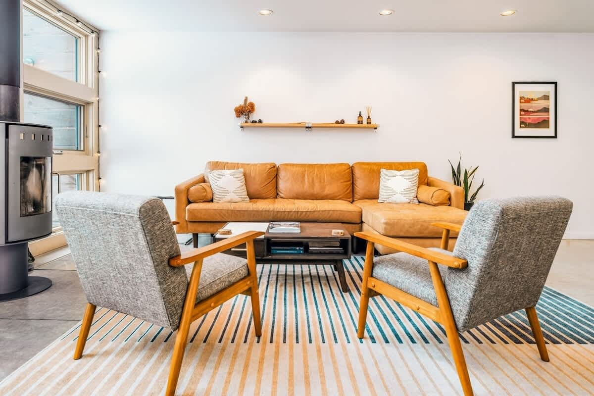 Inviting living room with a tan leather sectional sofa and two grey armchairs arranged on a striped rug, facing a dark wood coffee table. A modern wood-burning stove is visible on the left, and a floating shelf with decor adorns the white wall.