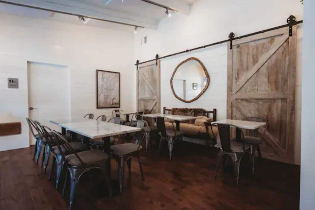 Bright and airy dining space with white walls, dark wood floors, several square marble-top tables with metal chairs, and two large rustic barn doors on a sliding track.