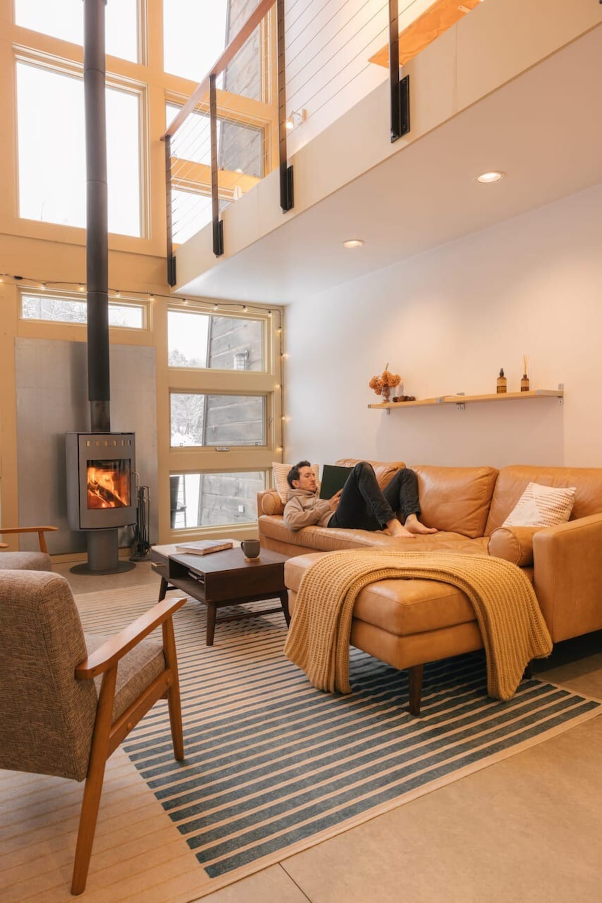 A man relaxes on a tan leather sectional sofa in a bright living room with a wood-burning stove and large windows. A striped rug, a dark wood coffee table, and a light gray armchair complete the inviting space, with a mezzanine visible above.