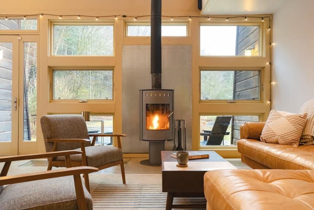 Cozy living room with a modern wood-burning stove as the focal point, surrounded by large windows offering views of a snowy, wooded landscape. A tan leather sofa and two grey armchairs are arranged around a dark coffee table, with string lights adding a warm ambiance.
