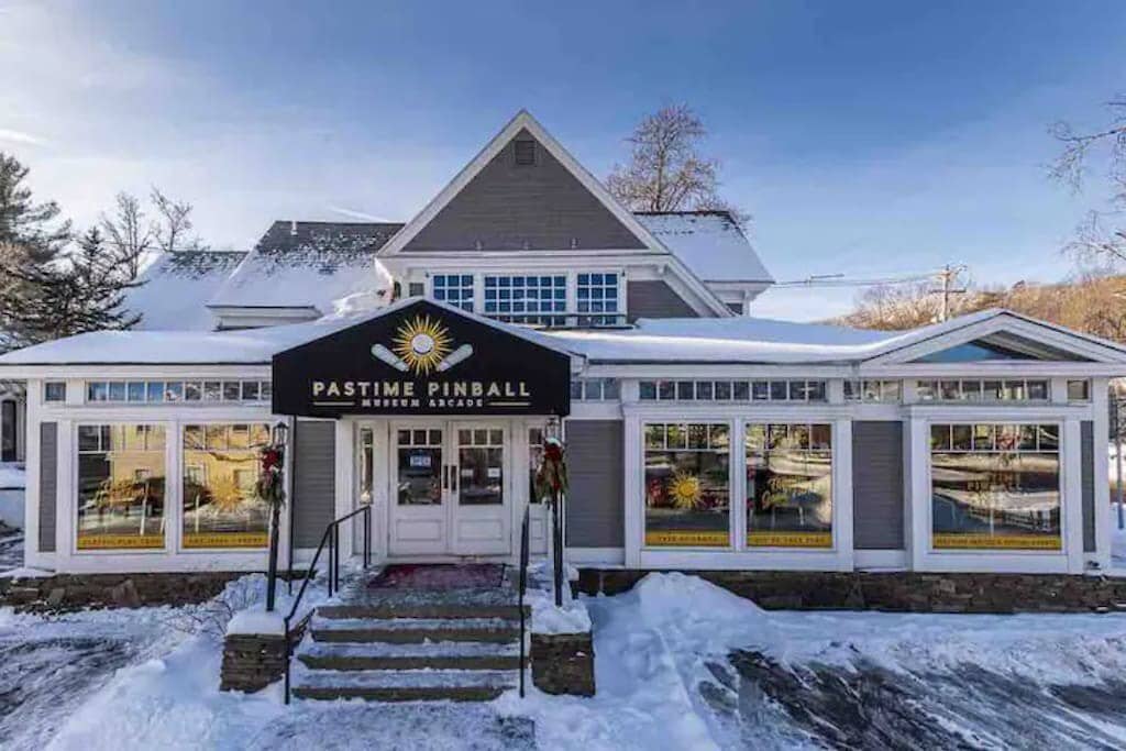 Charming "PASTIME PINBALL MUSEUM ARCADE" building with a gabled roof and large windows, covered in a blanket of snow under a clear blue sky, featuring a prominent black awning.