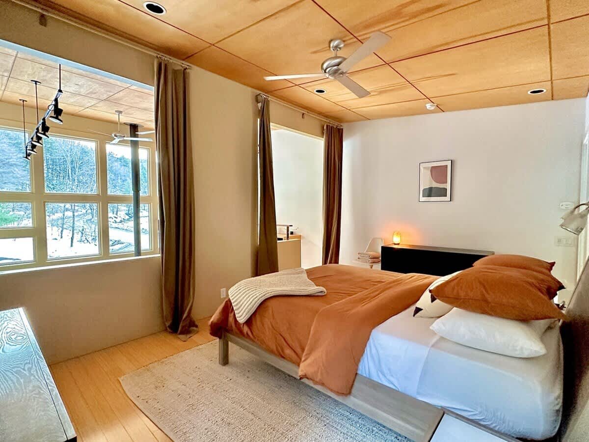 Spacious bedroom featuring a bed with rust-colored bedding, large windows with brown curtains, and a light wood plank ceiling. A ceiling fan hangs centrally, and a modern abstract artwork adorns the wall.