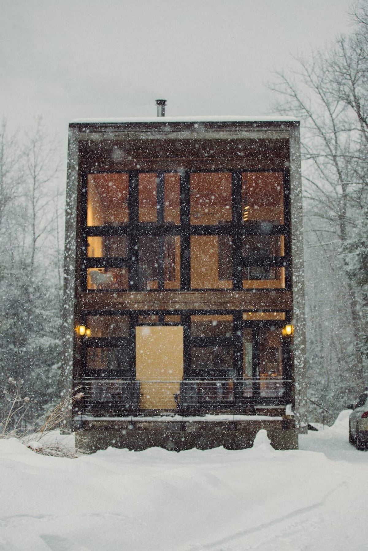 Modern multi-story house with large windows and dark siding, covered in a fresh blanket of snow during winter. The structure features clean lines and a minimalist aesthetic, blending with the snowy natural surroundings.