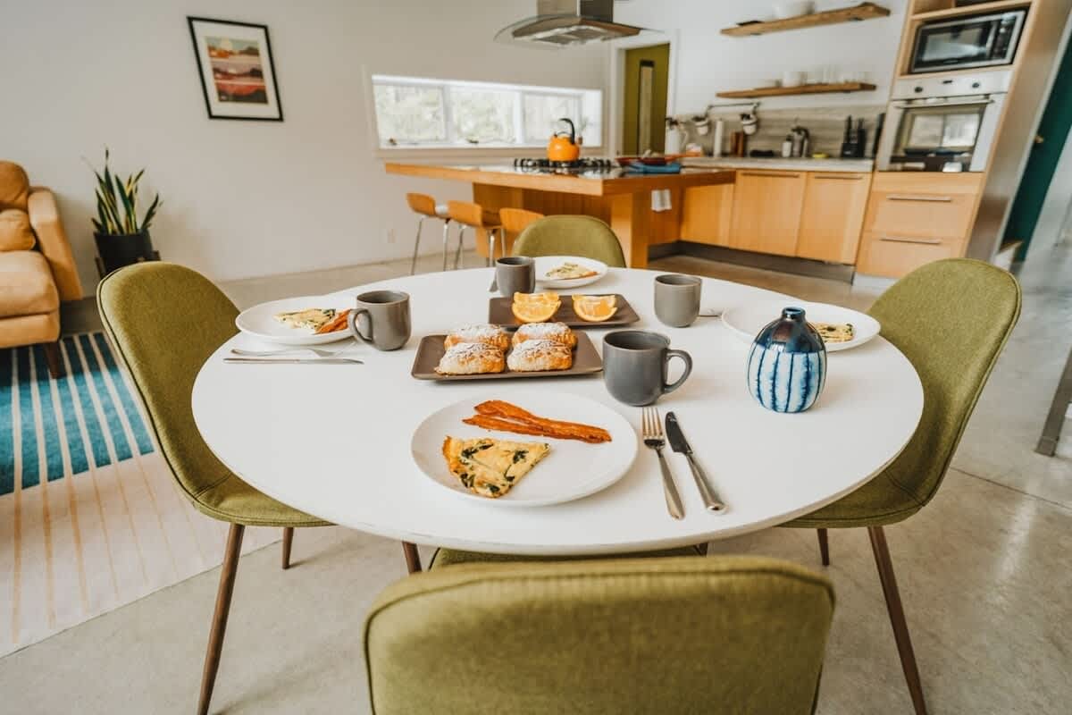 A bright, modern dining area features a round white table set for four with breakfast items, including pastries, bacon, and eggs. The open-concept space connects to a kitchen with light wood cabinetry and a large island.