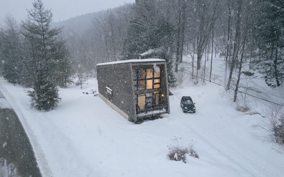 Modern rectangular cabin with dark siding and large windows, nestled in a snow-covered forest with bare trees, a car parked nearby, and smoke gently rising from a chimney.