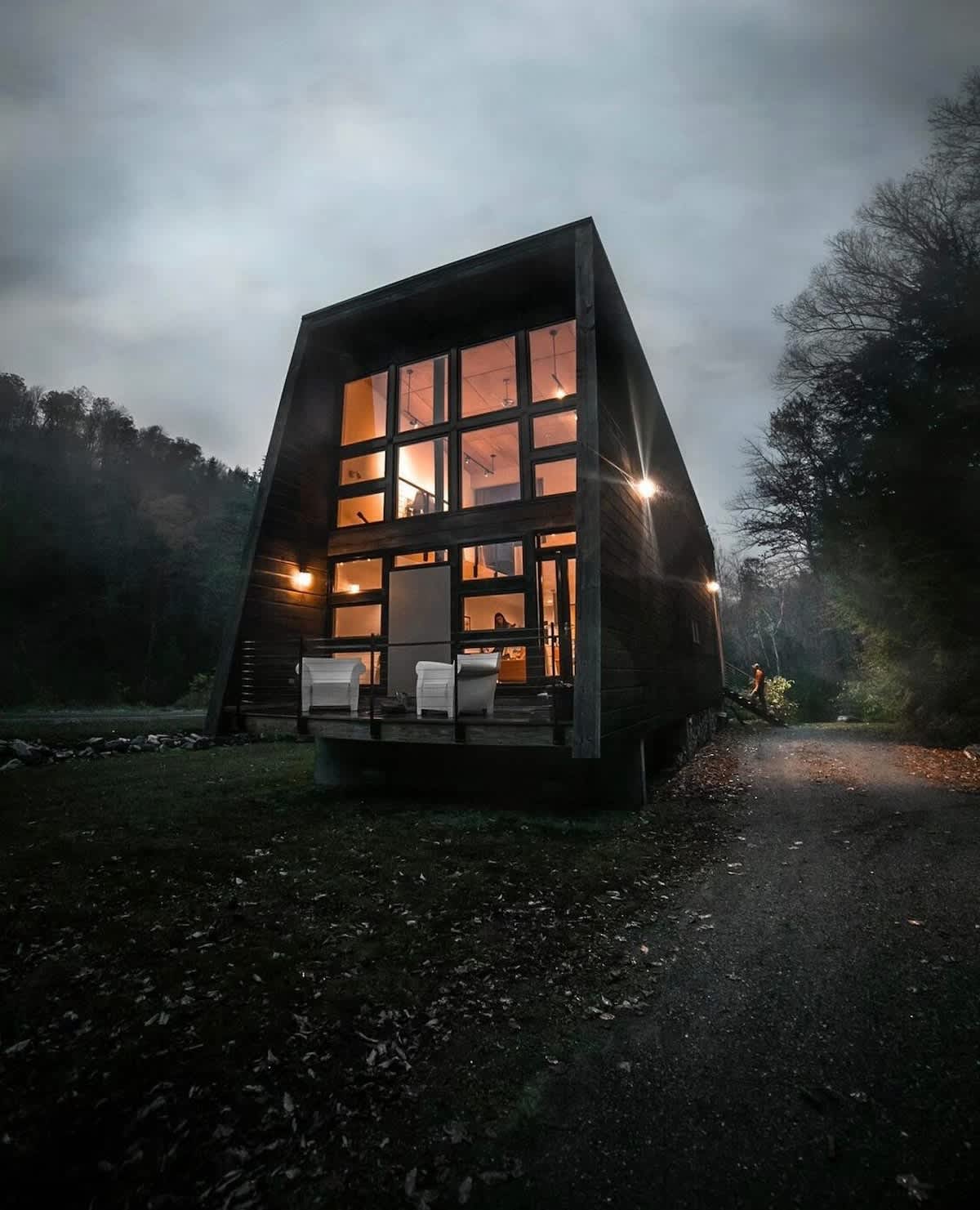 The modern cabin glows warmly at dusk, with its interior lights illuminating the expansive, multi-pane windows that dominate the front facade. The dark wood siding and angled roofline stand out against the darkening, tree-lined backdrop.