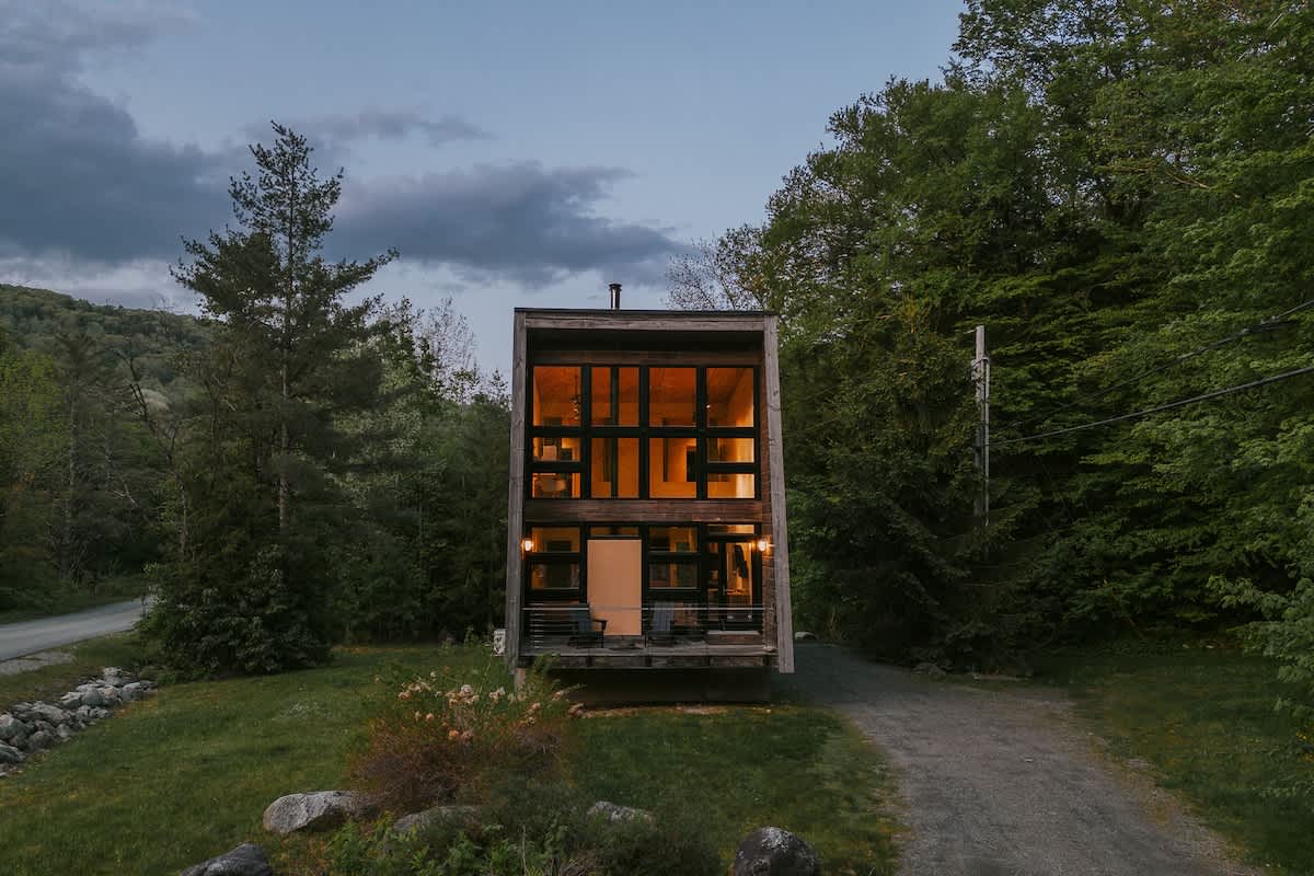 The tall, narrow modern cabin stands prominently with its full-height, multi-pane windows and dark wood siding, showcasing its distinctive angled architecture. A small deck with outdoor seating is visible at the entrance, surrounded by lush trees and a gravel driveway.