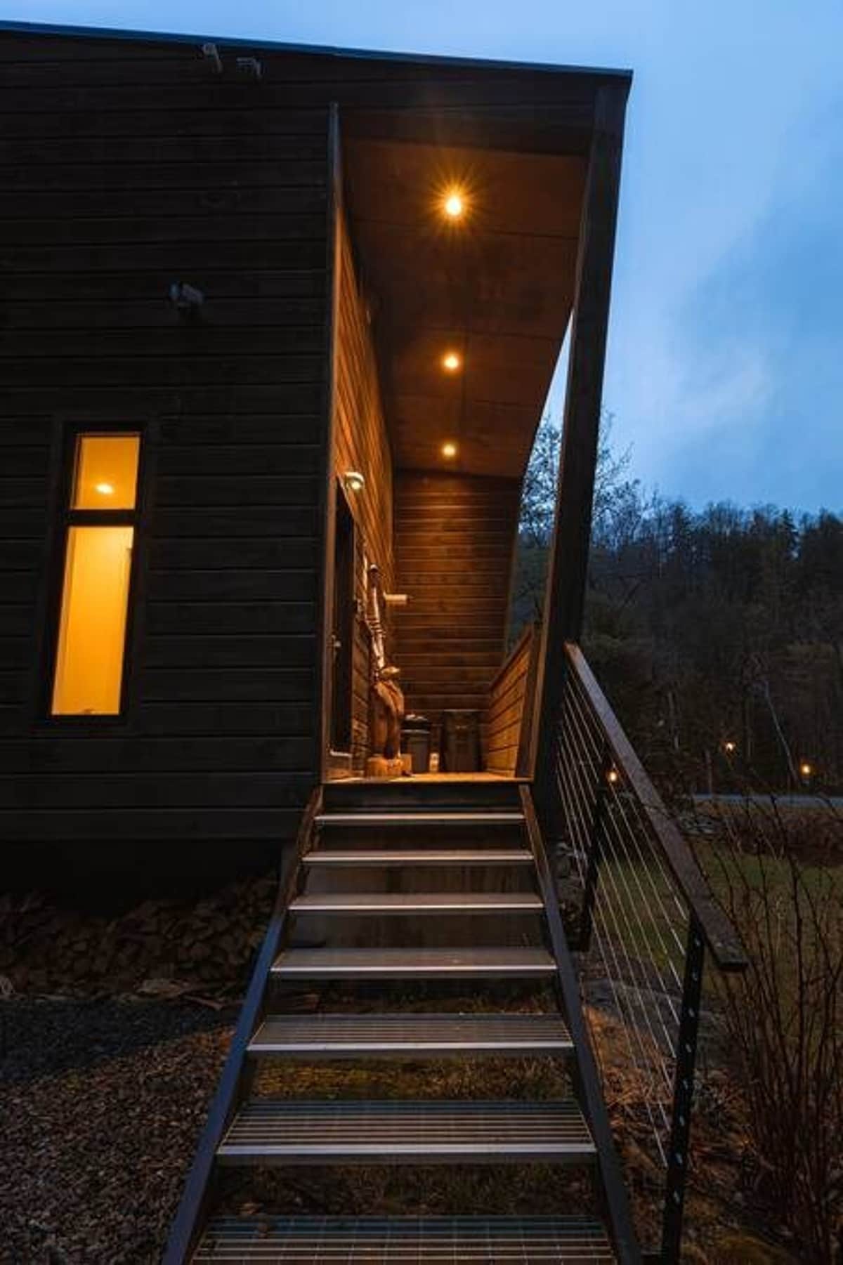 The modern entrance stairs, made of metal with cable railings, are warmly illuminated by recessed ceiling lights at dusk, leading up to the dark wood-sided cabin. The surrounding area is dark, emphasizing the inviting glow of the entrance.