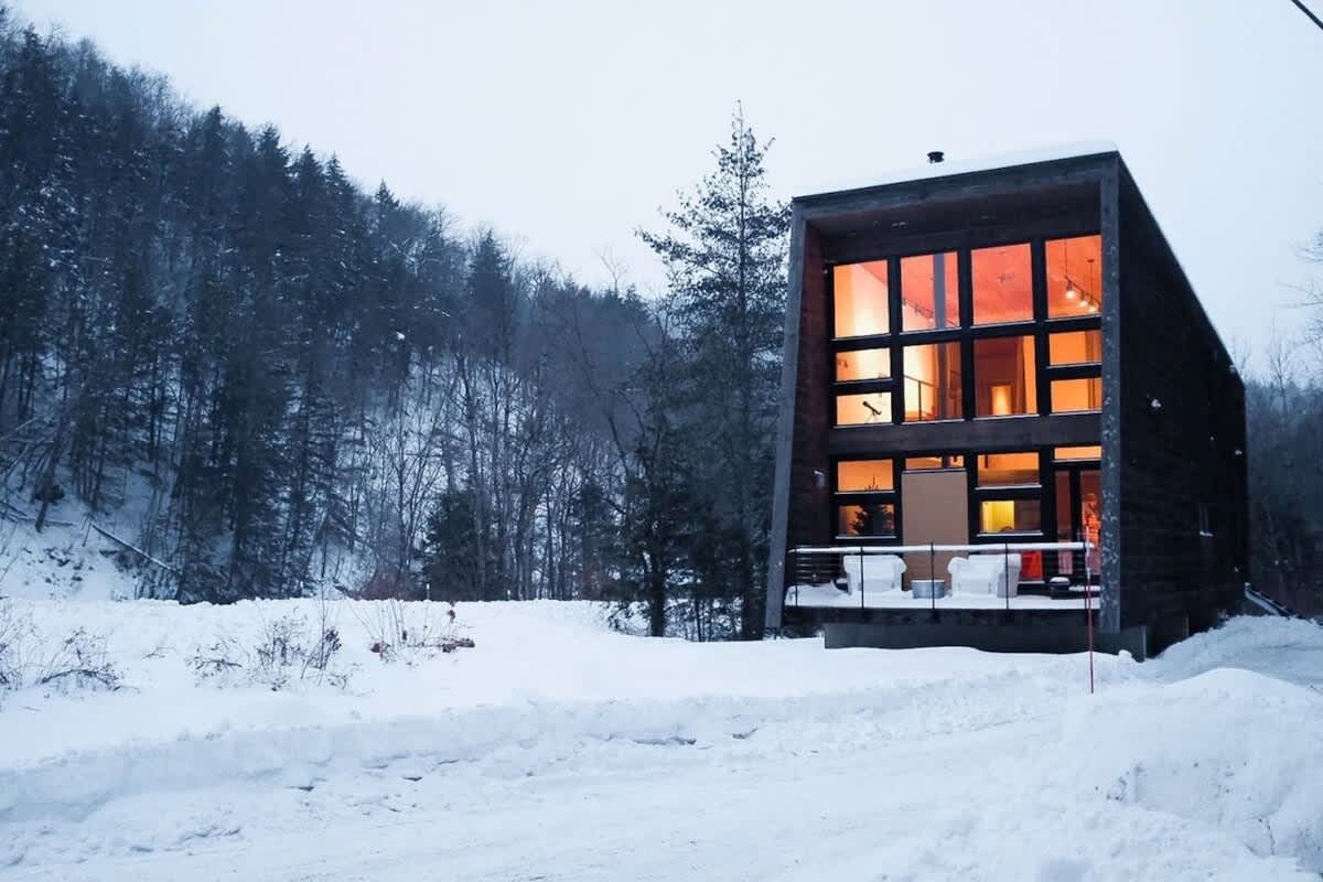 Modern, dark-clad cabin with large, multi-pane windows showcasing warm interior lighting, set against a snowy, wooded mountain backdrop. The house has a distinctive angular design and a small outdoor seating area on a deck.