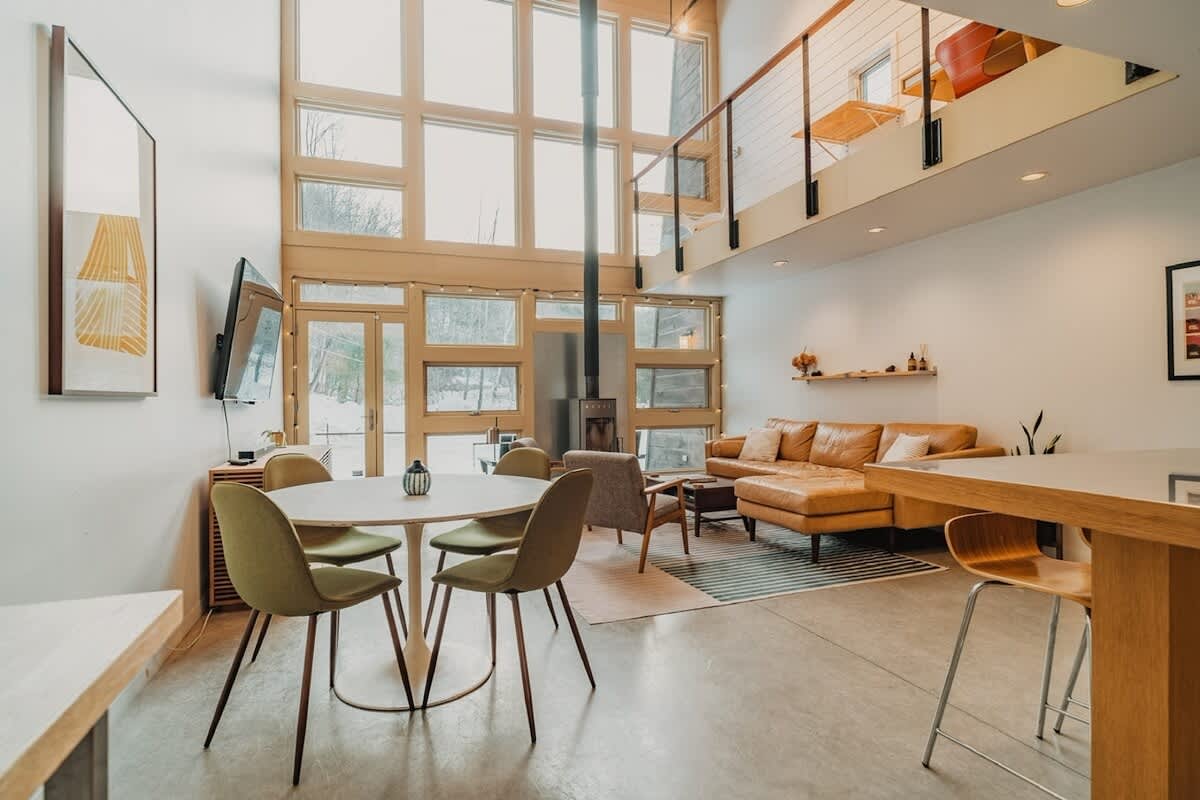 Spacious open-concept living area with high ceilings and floor-to-ceiling windows offering views of a snowy landscape. A tan leather sectional sofa and a modern dining table with green chairs define the space, featuring light concrete floors.