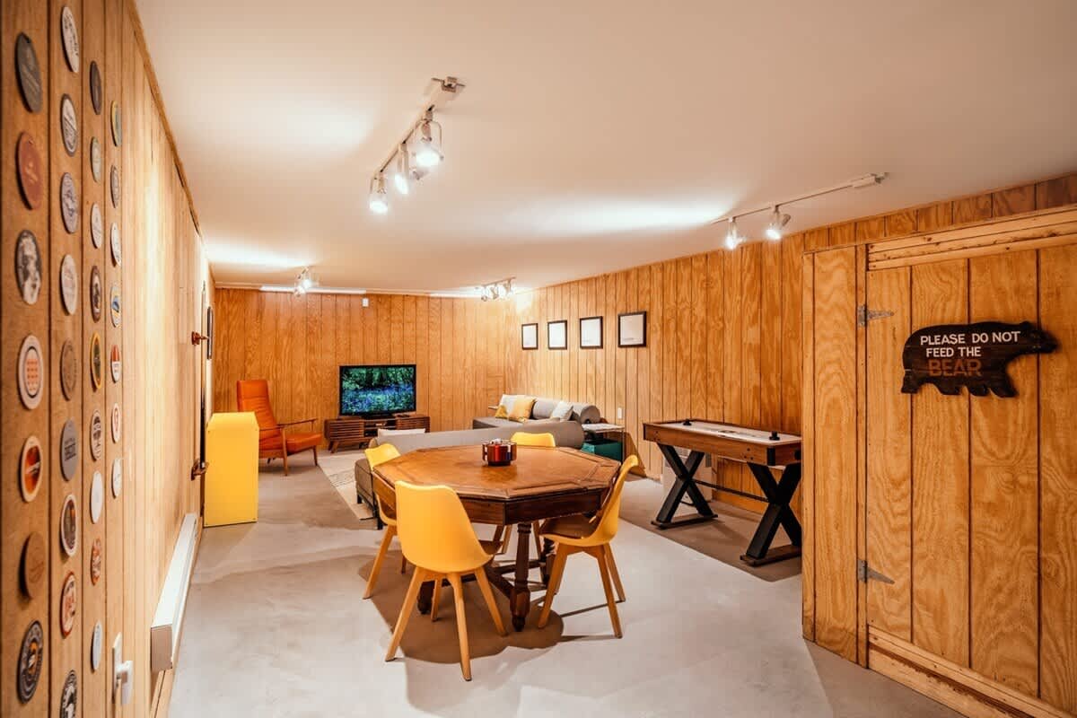 A retro-inspired game room with wood-paneled walls and concrete floors. It features an octagonal wooden dining table with yellow chairs, a shuffleboard table, a TV area with a gray sofa, and a "Please Do Not Feed The Bear" sign on a wooden door.