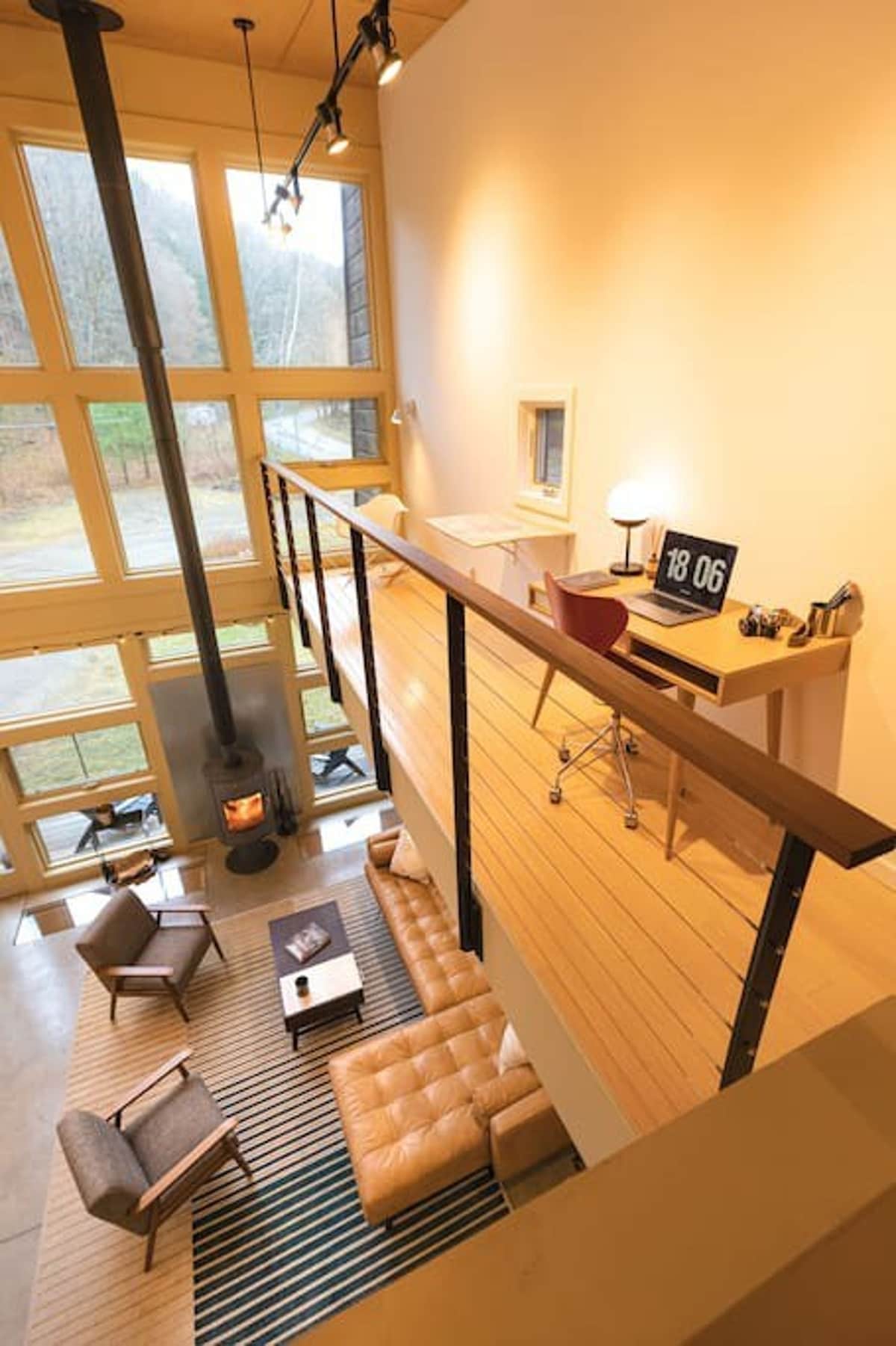 An elevated view of the living room below, showcasing a modern office nook on a wooden mezzanine. The office features a light wood desk with a laptop, a red rolling chair, and a minimalist lamp, overlooking the double-height living space with a wood-burning stove and large windows.