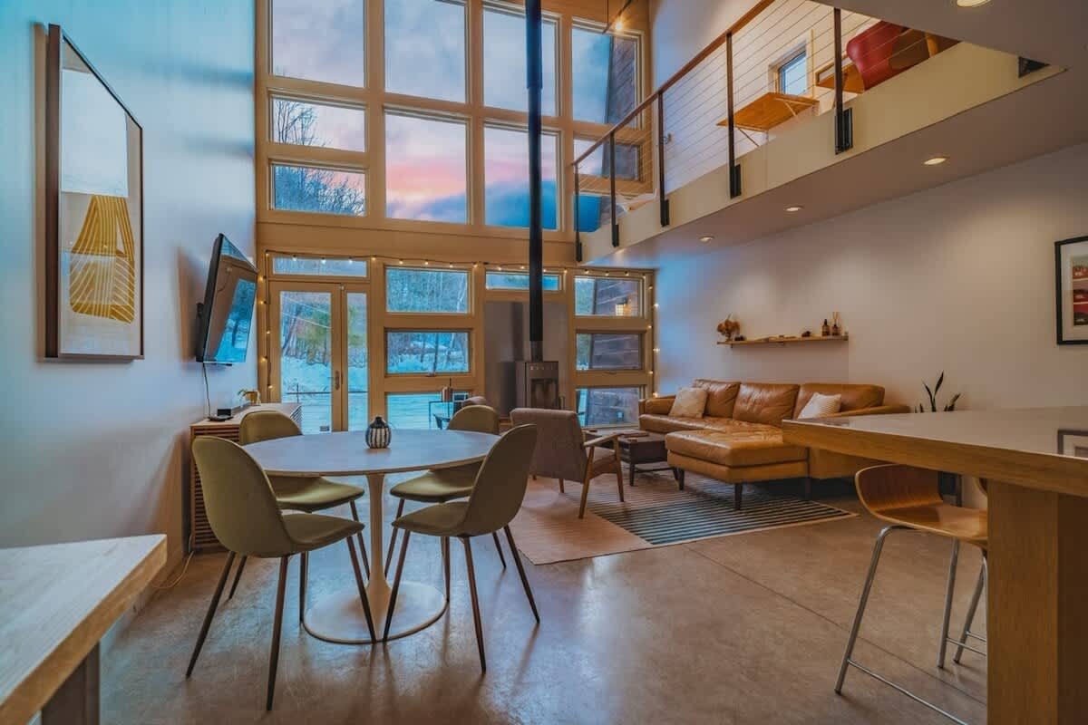 A wide shot of the open-concept living and dining area, featuring a tan sectional sofa, a round dining table with olive green chairs, and a wood-burning stove. Large, multi-pane windows offer panoramic views of a winter landscape and a colorful sunset.