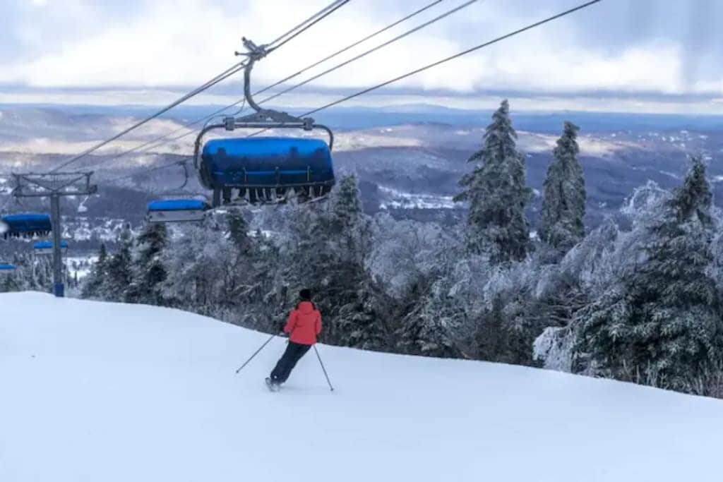 A skier in a red jacket descends a pristine snow-covered slope, with a blue ski lift carrying passengers overhead. Frost-covered evergreen trees and distant mountains complete the winter landscape.