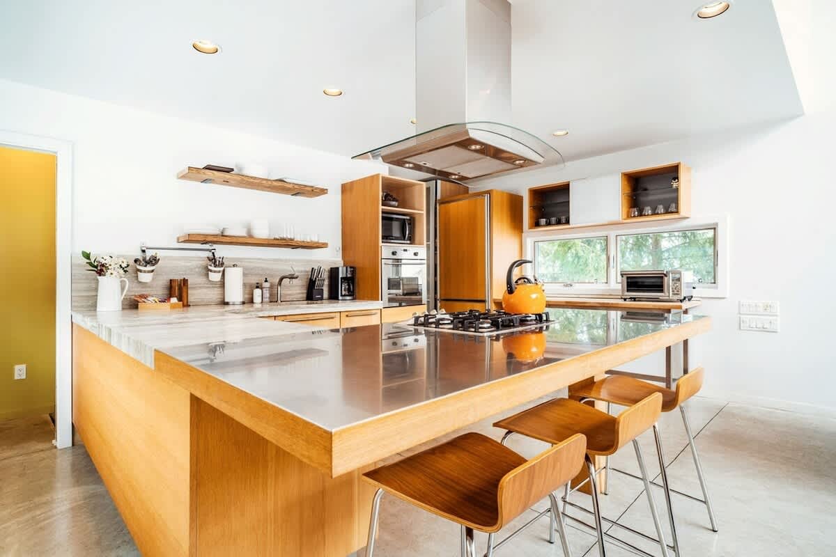 Modern kitchen with light wood cabinetry, a large island featuring a stainless steel countertop and integrated gas cooktop, and three wooden bar stools. Overhead, a sleek range hood with a glass canopy is visible, and the space is brightly lit with recessed lighting.