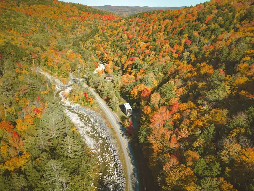 A vibrant aerial view showcases the modern cabin surrounded by a breathtaking autumn landscape, with a river winding through a valley of trees ablaze in red, orange, and yellow fall foliage. The house is nestled amidst the colorful forest.