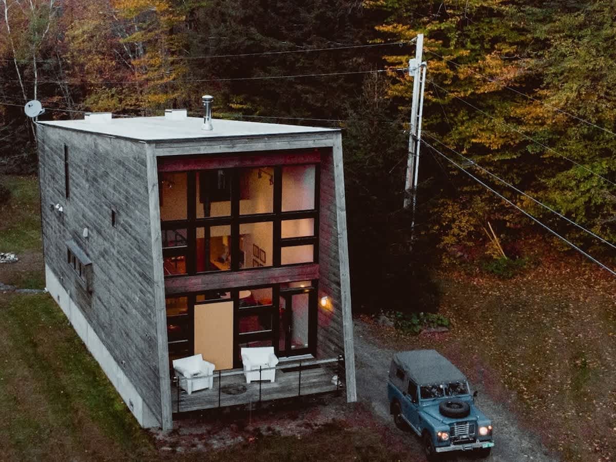 Aerial view of the modern cabin with dark wood siding and a flat roof, showing a gravel driveway with a vintage car, and surrounded by dense autumn trees.