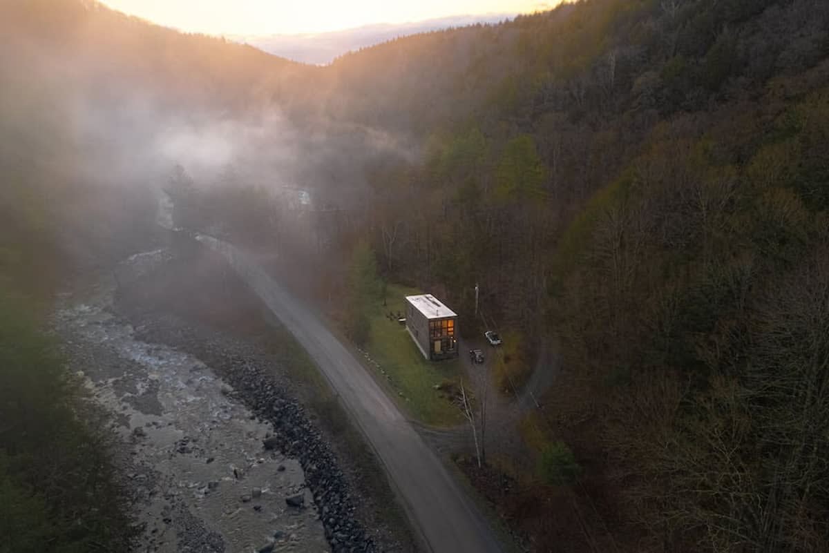 An aerial view captures the modern cabin nestled in a misty, tree-filled valley alongside a winding river, with the warm glow of sunrise or sunset illuminating the distant horizon. The house appears secluded and surrounded by nature.