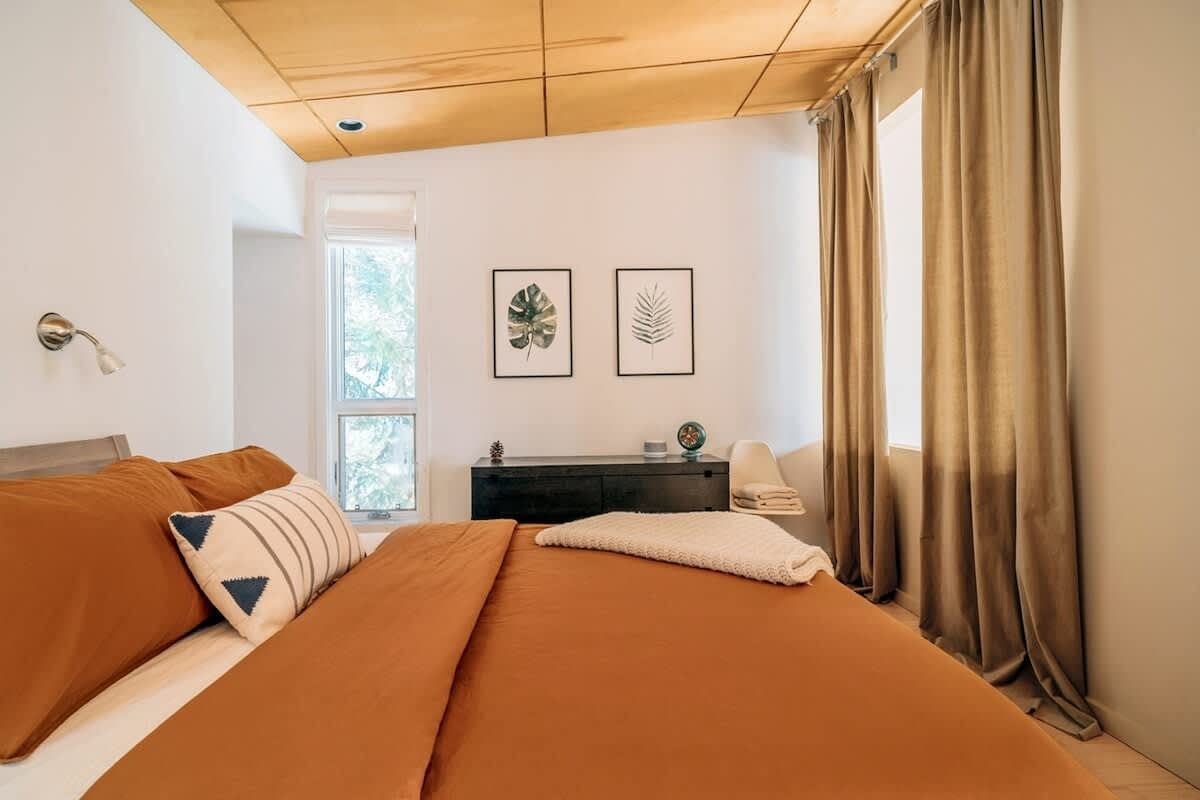 Cozy bedroom with a bed featuring rust-colored bedding and a striped accent pillow. Two framed botanical prints hang above a dark dresser, and a window provides natural light, all set against light hardwood floors.