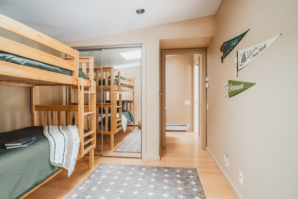 View of the bunk bed room showing the wooden bunk beds, a mirrored closet, and a doorway leading to a hallway. Decorative pennants adorn the wall, and a patterned rug covers the light hardwood floor.