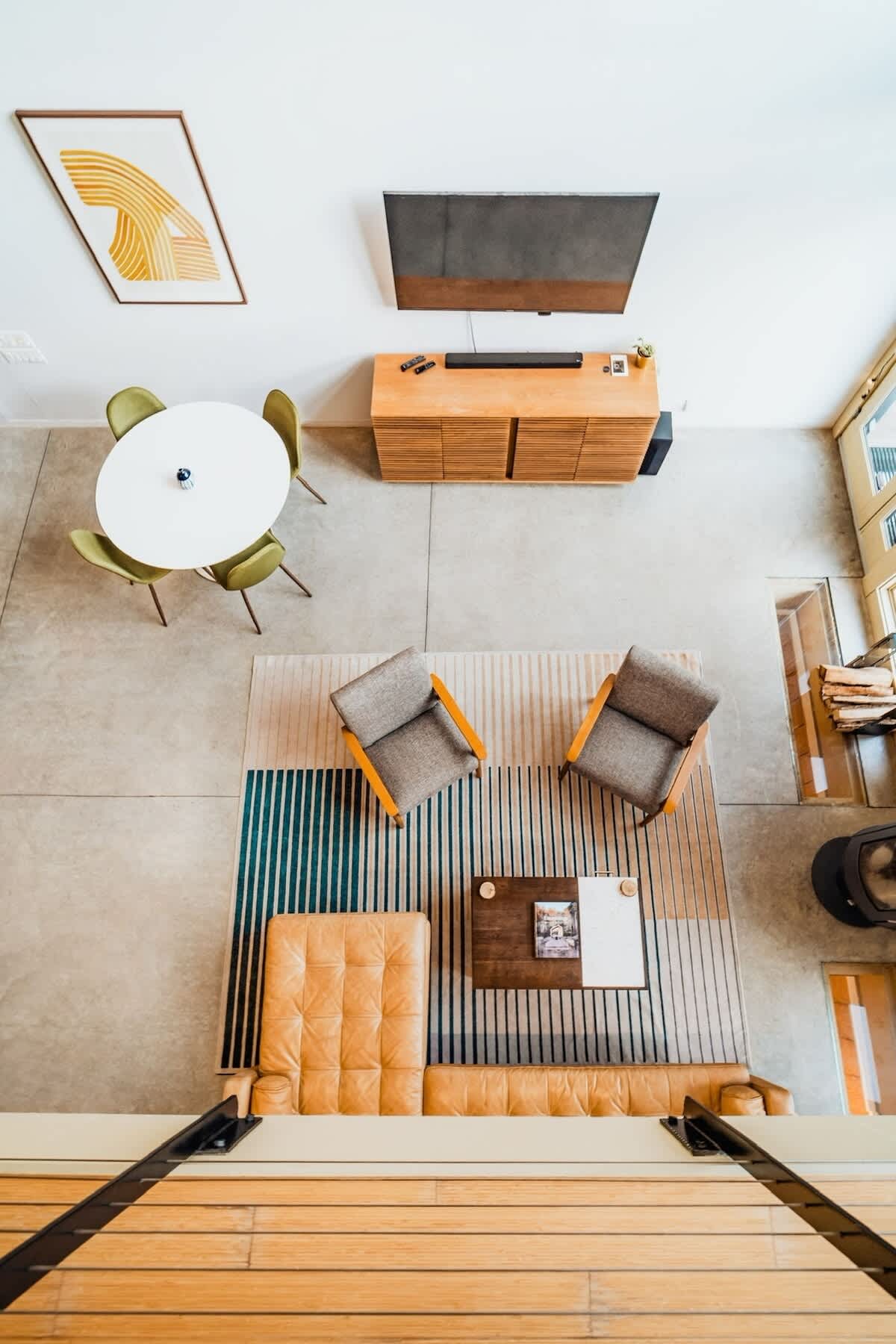 Aerial view of an open-concept living and dining area with concrete floors. The living space features a tan leather sectional, two armchairs, and a coffee table on a striped rug, while a round white dining table with four green chairs is visible nearby.