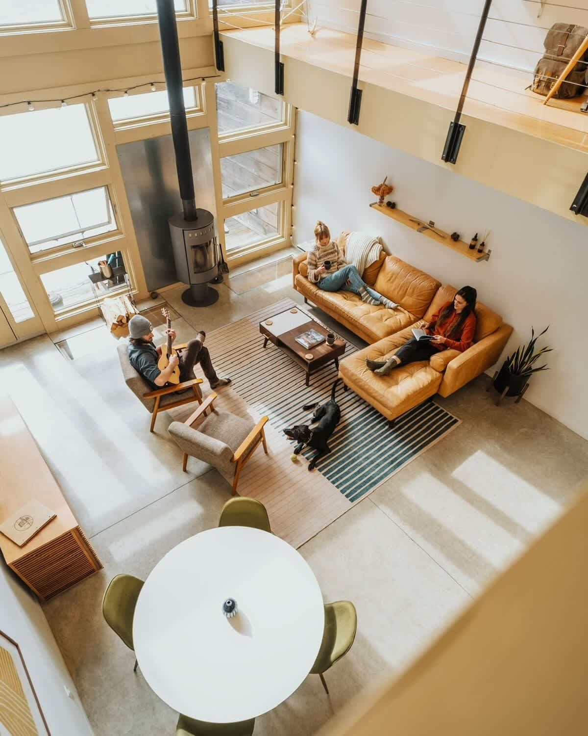 An aerial view of a spacious, open-concept living area with a high ceiling and large windows. A tan sectional sofa, two armchairs, a coffee table, and a striped rug define the seating area, with a wood-burning stove and dining table nearby. People are relaxing, and a black dog is on the rug.