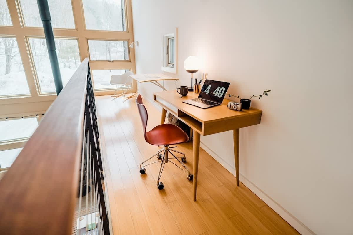 A bright, minimalist office space on a wooden mezzanine, featuring a light wood desk with a laptop displaying "18 06", a red rolling chair, and a modern globe lamp. Large windows offer a view of snowy trees outside, and a white rocking chair is visible in the background.