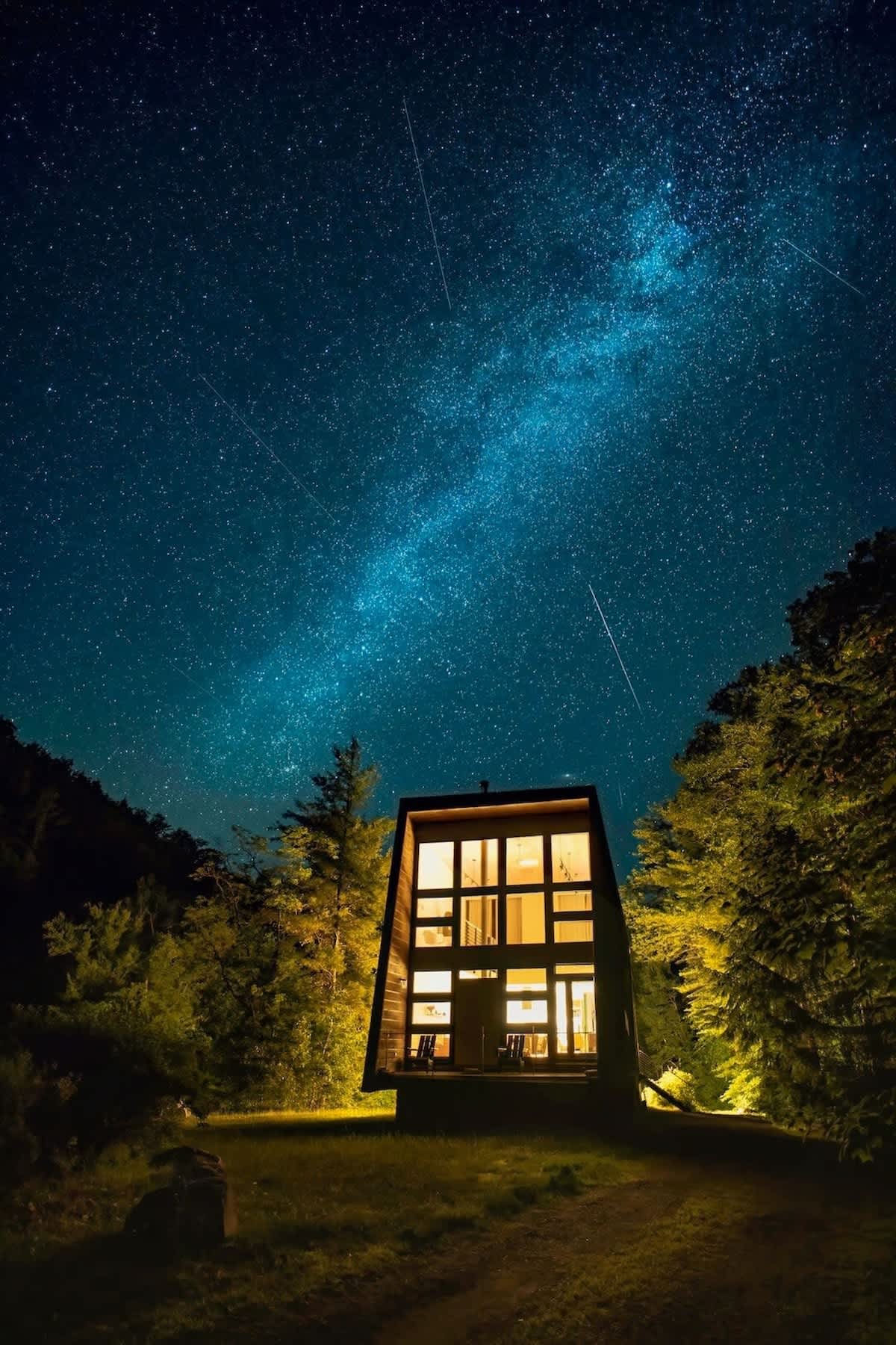 A striking modern cabin with large illuminated windows stands nestled in a dense forest under a breathtaking starry night sky, showcasing the Milky Way. The dark exterior contrasts with the warm interior light, creating a serene and isolated ambiance.