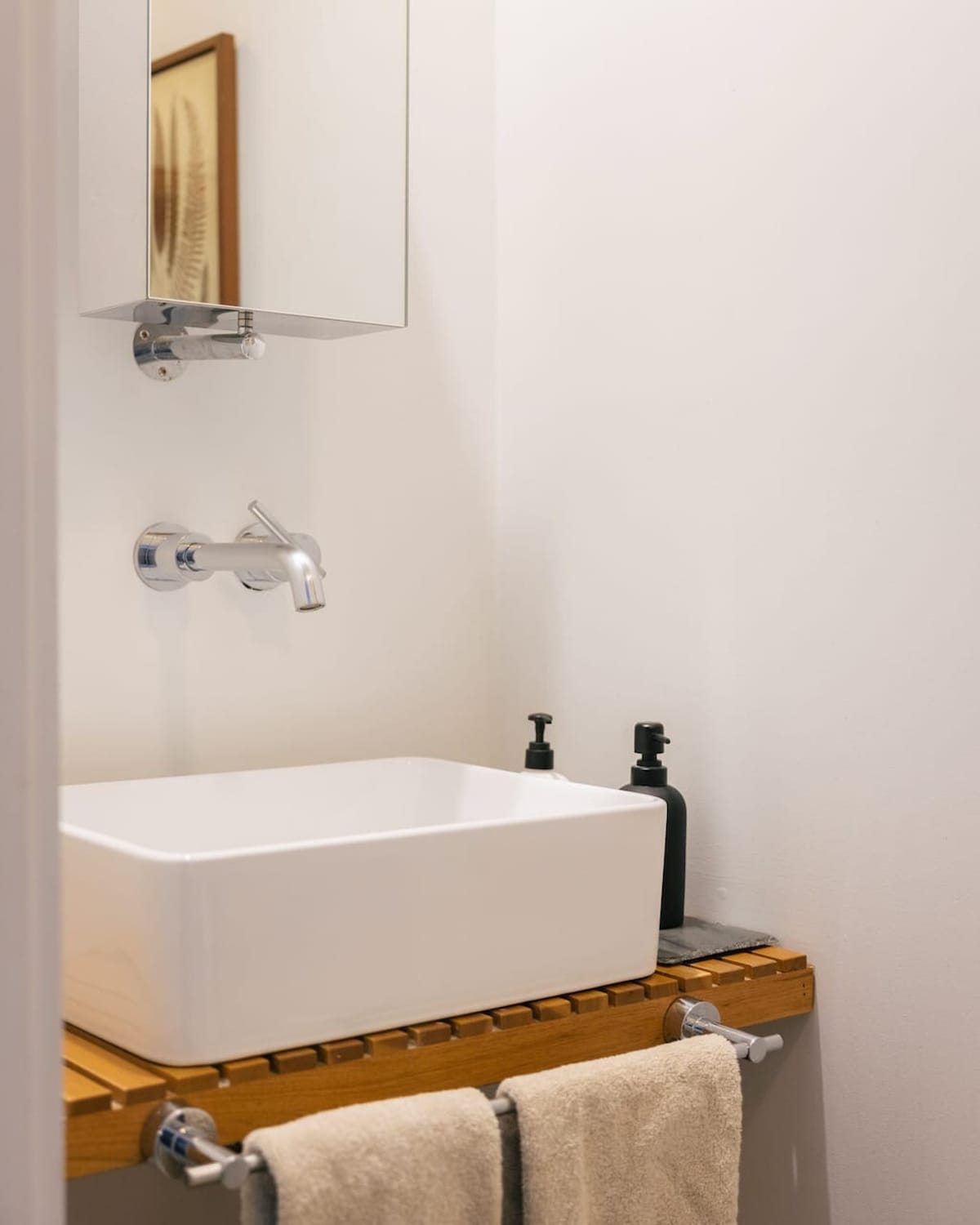 Modern bathroom sink area with a rectangular vessel sink on a slatted wooden vanity, chrome wall-mounted faucet, and a mirrored medicine cabinet above. Two dark soap dispensers and hand towels are neatly arranged.