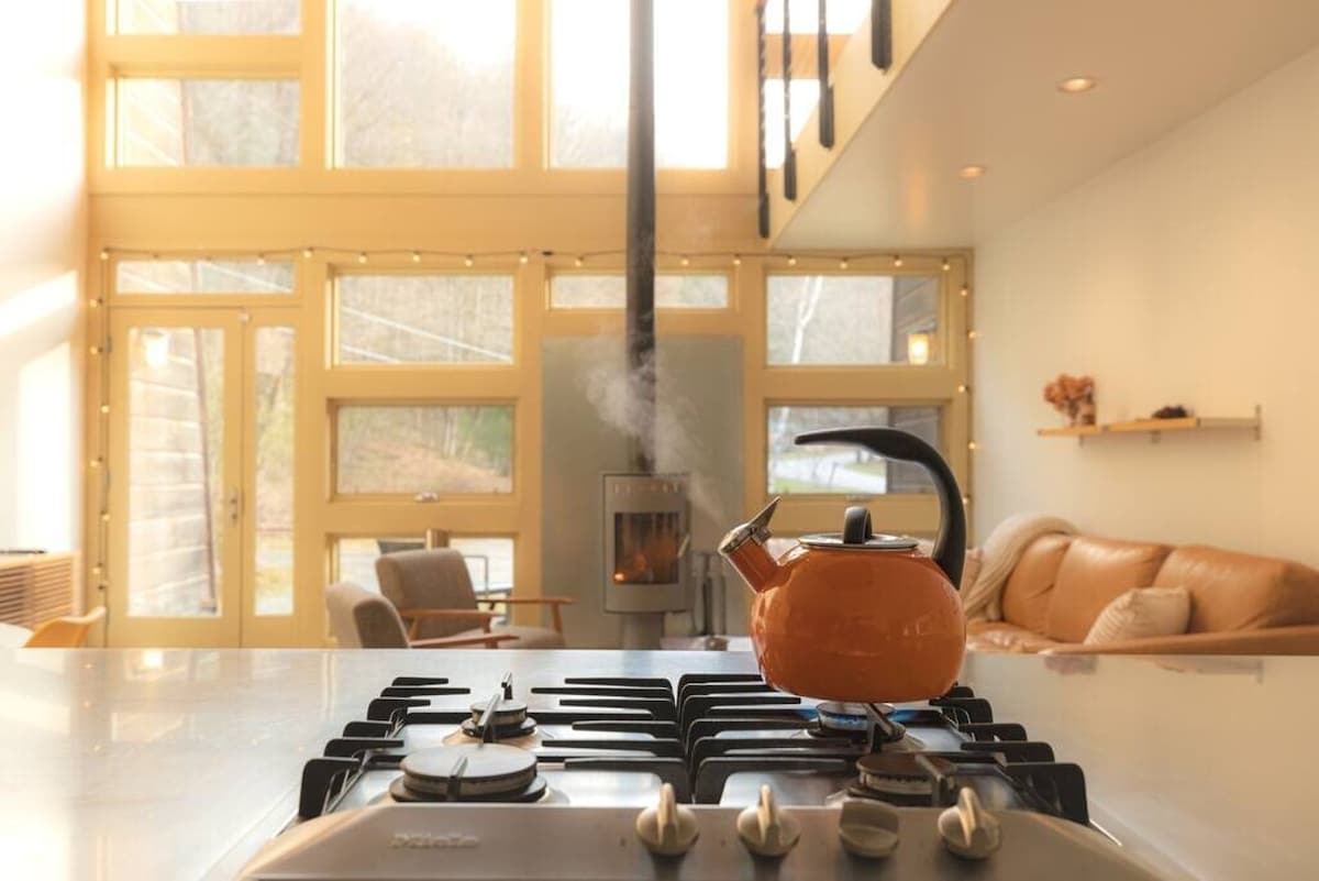 A vibrant orange kettle steams on a modern gas stovetop in a sunlit kitchen, with large windows revealing a snowy, wooded landscape outside. A cozy living area with a leather sofa and wood-burning stove is visible in the background.