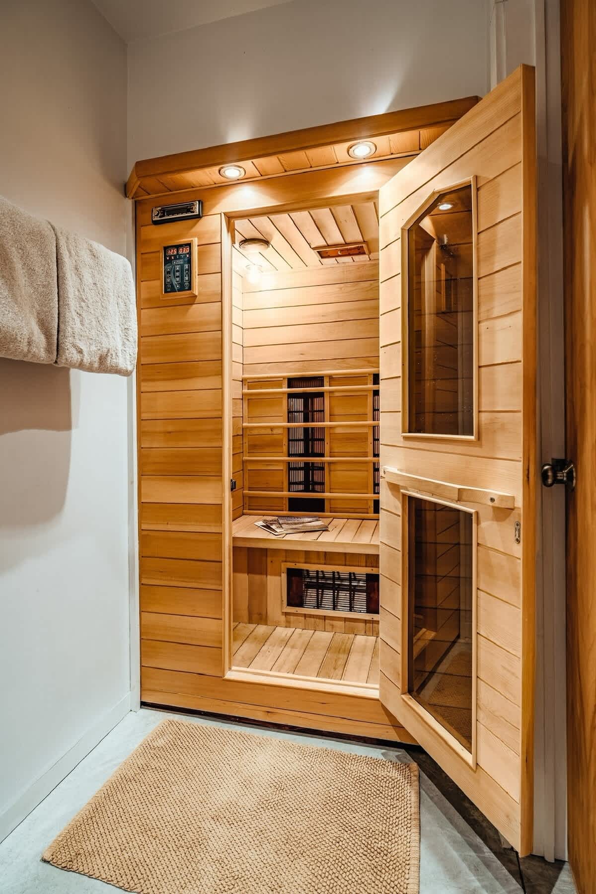 A compact, light wood sauna with an open door, revealing slatted benches and infrared heating panels. The interior is well-lit with recessed lighting, and a control panel is visible on the wall, indicating it's ready for use.