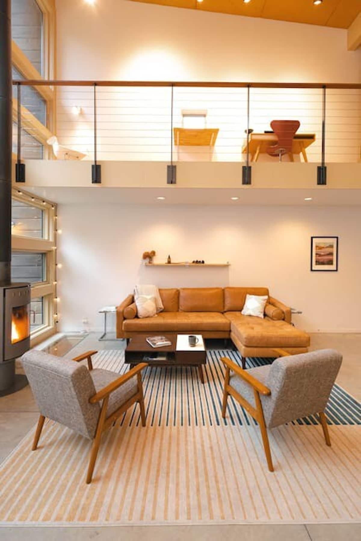 Bright living room with a high ceiling and an open loft area visible above, featuring a modern wood-burning stove and large windows. A tan leather sectional and two armchairs are arranged on a striped rug, with a wooden railing separating the loft.