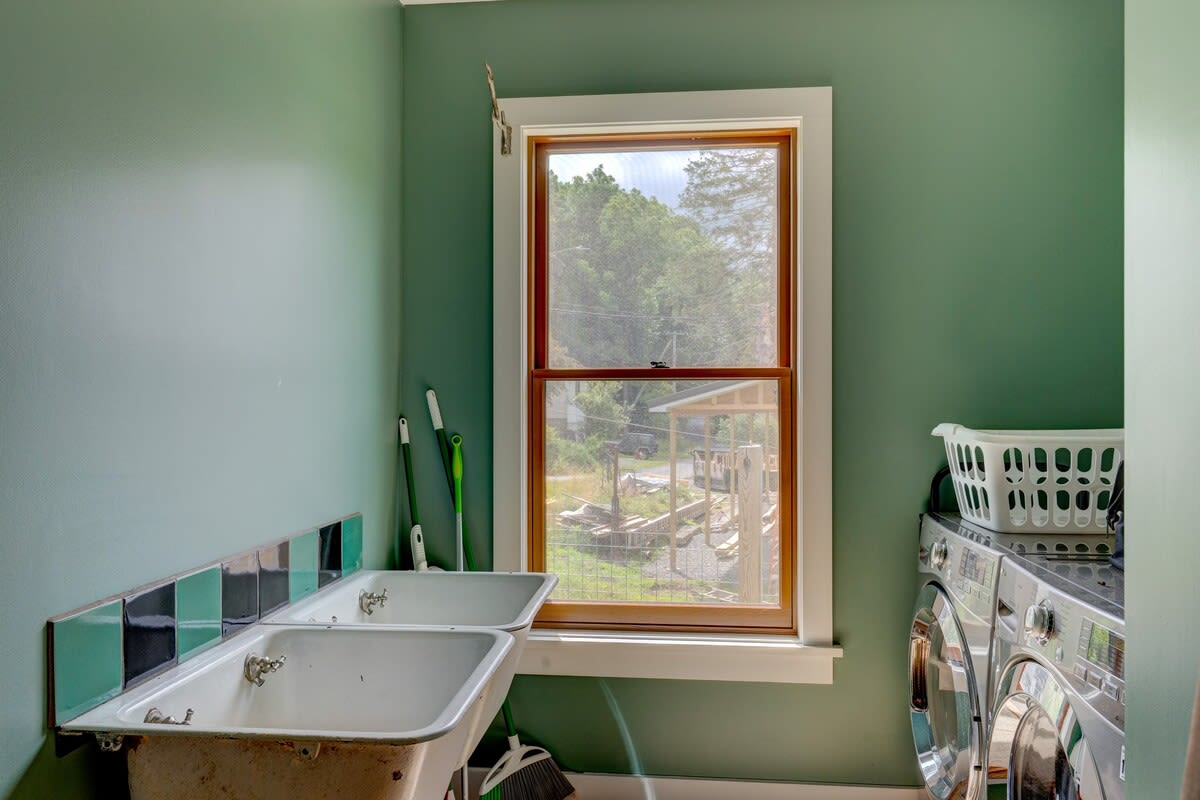 A functional laundry room with sage green walls, featuring two vintage-style utility sinks with black and green tiled backsplash, and a stacked washer and dryer unit. A large window overlooks a wooded area.