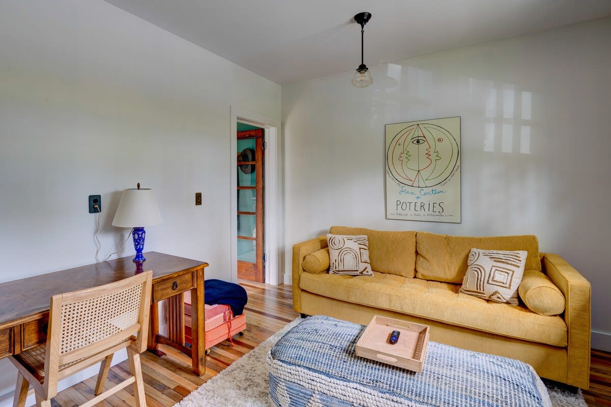 A cozy living room with a mustard yellow sofa adorned with patterned pillows, a blue and white woven ottoman, and a wooden desk with a cane chair. A large abstract art print hangs on the white wall.