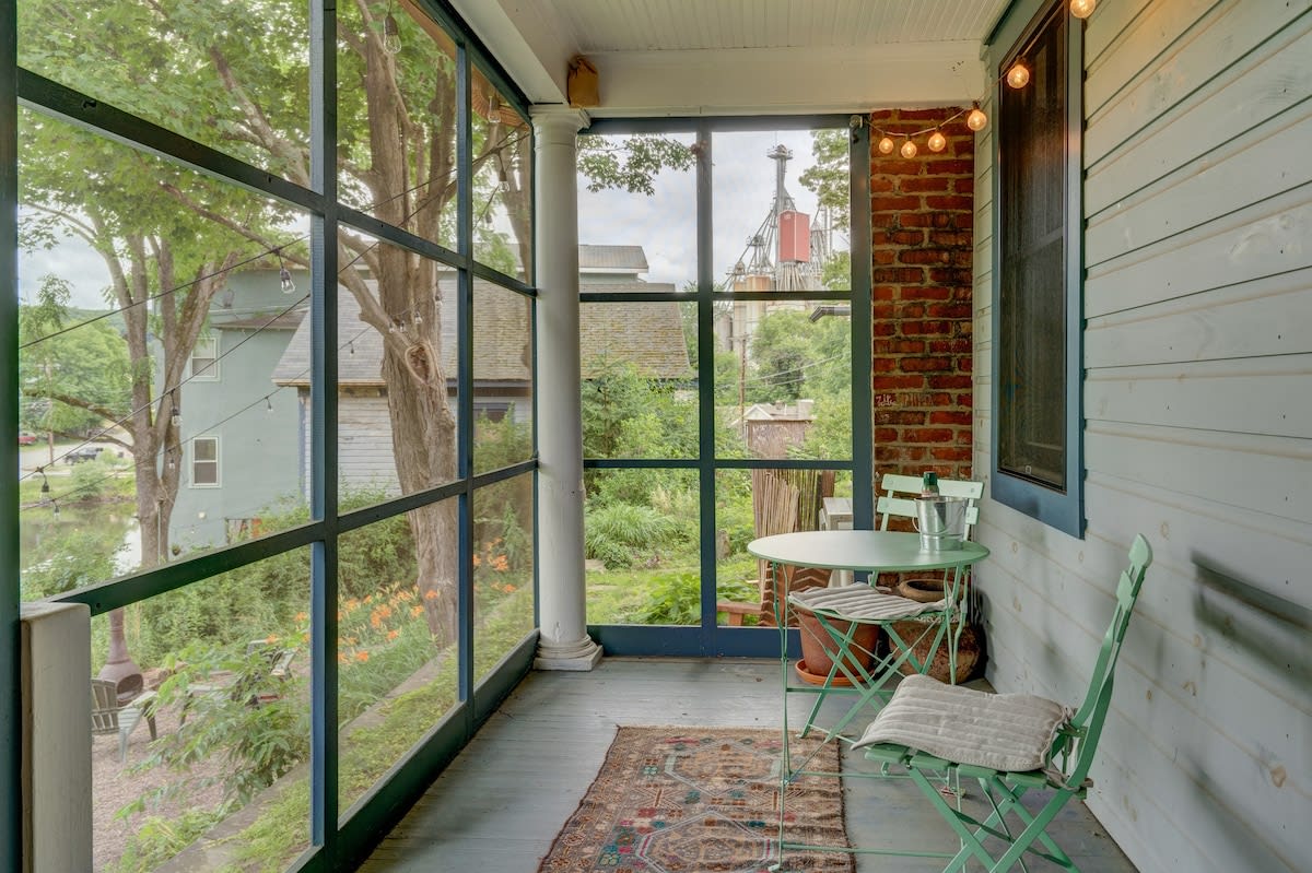 A screened-in porch with light blue painted floors, a small mint green bistro table and chairs, string lights, and views of trees and a nearby building through large windows.
