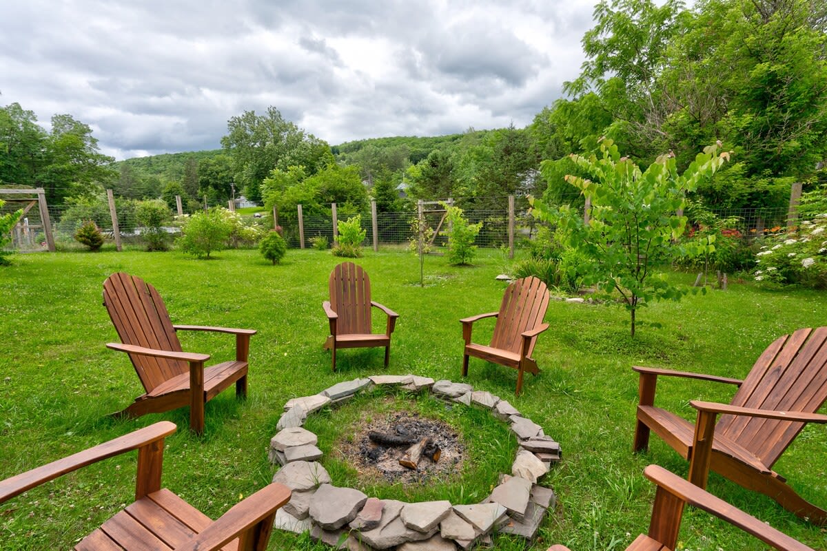 A stone fire pit surrounded by several wooden Adirondack chairs on a grassy lawn, with lush green trees and a cloudy sky in the background, suggesting a spacious backyard.