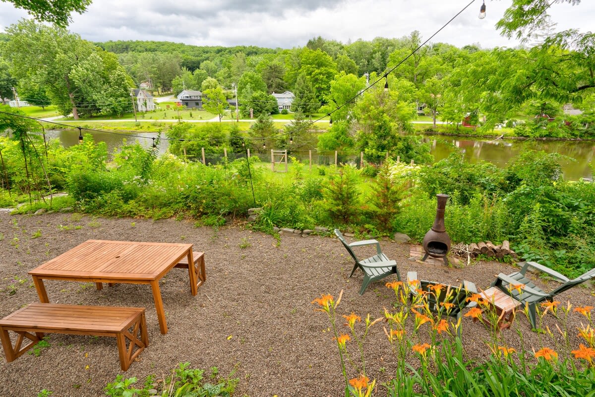 A spacious gravel yard area featuring a wooden picnic table with benches, a kiva-style fire pit with Adirondack chairs, and vibrant orange daylilies, all overlooking a serene lake and lush green hills.